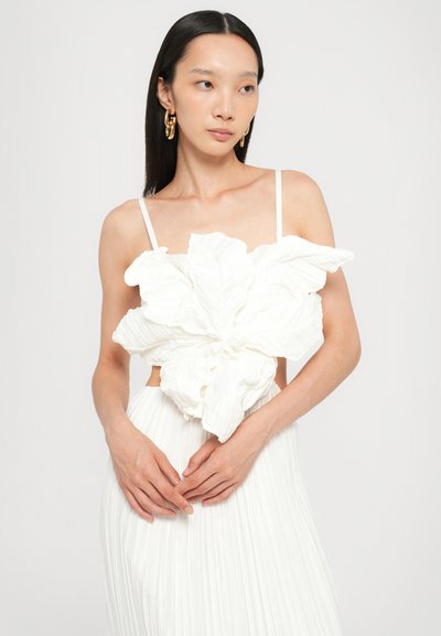 Hvid kjole med tekstureret, oversized blomsterdetalje ved brystet; har tynde stropper og plisseret nederdel. Model poser med hænderne foldet.