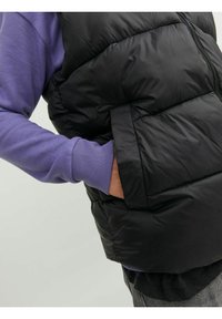 Black puffer vest with a textured surface, featuring a side pocket. Worn over a purple long-sleeve shirt with ribbed cuffs.