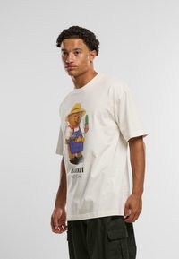 Creme bomulds t-shirt med et tegneserie bjørn i en forklæde, der holder en kaktus, med teksten "MARKED Selvpleje" under designet. Korte ærmer.