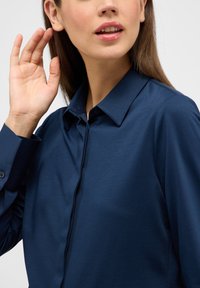 Camicia blu navy con colletto, caratterizzata da una texture liscia e maniche lunghe. Dettagli notevoli includono i bottoni e un design pulito.