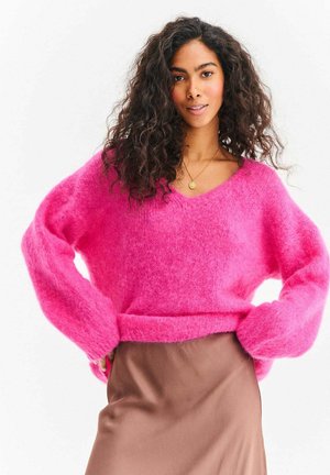 Pluszowy różowy sweter z dekoltem w serek i oversize'owymi rękawami, zestawiony z brązową satynową spódnicą. Miękka tekstura i fason na zakładkę.