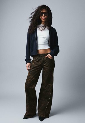 Göndör hajú nő napszemüvegben, sötétkék cipzáras kapucnis pulóvert, fehér crop topot, leopárdmintás bő szárú nadrágot és fekete cipőt viselve, szürke háttér előtt áll.