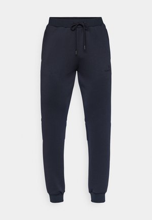 Calças de treino azul-marinho com cintura ajustável por cordão, punhos elásticos, dois bolsos laterais e uma marcação subtil na perna esquerda. Textura de tecido suave.