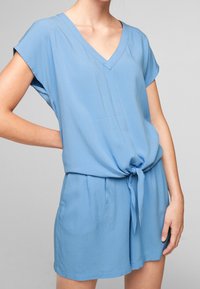 Jumpsuit court bleu clair avec décolleté en V, manches courtes et détail noué à la taille. Fabriqué en tissu texturé pour un intérêt supplémentaire.