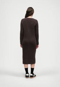 Μακρύ, εφαρμοστό καφέ ριπ φόρεμα με μακριά μανίκια. Το υλικό φαίνεται υφαντό. Φορεμένο με μαύρες κάλτσες και μαύρα-λευκά αθλητικά παπούτσια.