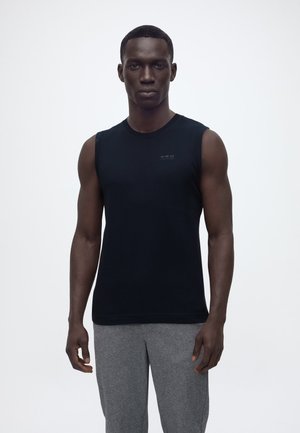 Mężczyzna stojący przodem, ubrany w czarny bezrękawnik i szare spodnie dresowe na jednolitym białym tle.