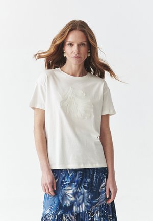 Witte katoenen t-shirt met een gestructureerd bladdesign op de voorkant, gecombineerd met een blauwe rok met bloemenpatroon; korte mouwen en ronde halslijn.