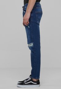 Plave traperice skinny kroja, s izbjeljenim detaljima na koljenu i istrošenom teksturom; uparene s crnim tenisicama.