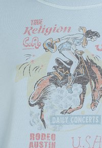 Jasnoniebieska koszulka z grafiką w stylu vintage przedstawiającą kobietę jadącą na koniu, z tekstem w kolorze czerwonym i żółtym, nawiązująca do tematyki rodeo.