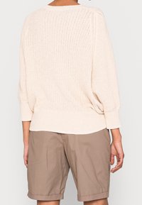 Suéter de punto beige claro con un corte relajado, mangas anchas y un dobladillo trasero redondeado, combinado con pantalones cortos caqui.