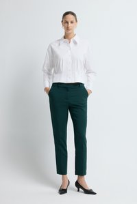 Camicia bianca corta abbottonata con colletto, abbinata a pantaloni verde scuro su misura. Modella in piedi di fronte con le mani nelle tasche, indossando tacchi neri.