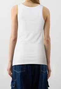 Mujer vestida con una blusa sin mangas blanca y pantalones cargo de mezclilla azul oscuro, mostrada desde atrás contra un fondo claro y liso.