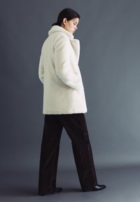 Kobieta w białym futrzanym płaszczu ze sztucznego futra, czarne szerokie spodnie i czarne buty, stojąca na tle szarego tła, trzymająca ręce w kieszeniach płaszcza.