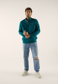 Blågrön zip-up tröja med hög krage, bärs med ljusblå slitna jeans och beige sneakers. Har en liten broderad logotyp på bröstet.