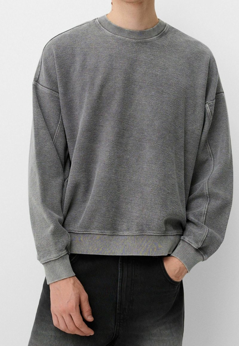 Personne portant un sweat-shirt gris à col rond ample avec des poignets et un bas côtelés, associé à un jean noir délavé.