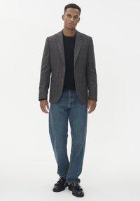 Šedý kostkovaný blazer s jedním knoflíkem, kombinovaný s tmavými džíny. Model má na sobě černý top a černé boty, stojící na bílém pozadí.