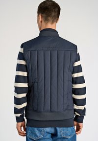 Marinblå pufferväst med vertikalt quiltad textur, med randiga långärmade tröjor i mörkblått och krämfärgat. Ribbad krage och midja.