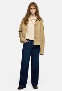 Beżowa zamszowa kurtka z dużymi guzikami, noszona na jasnokolorowej koszulce polo. Połączona z ciemnymi dżinsami o szerokich nogawkach i brązowymi butami.