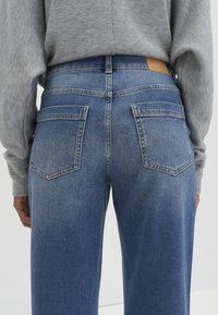 Personne portant un jean en denim bleu taille haute et un pull gris, vue de dos, mettant en valeur les poches arrière et les détails de la taille.
