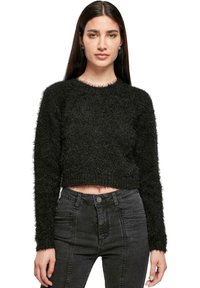 Donna che indossa un maglione corto nera e pelosa, con jeans grigi scuri a vita alta, in piedi davanti a uno sfondo bianco.
