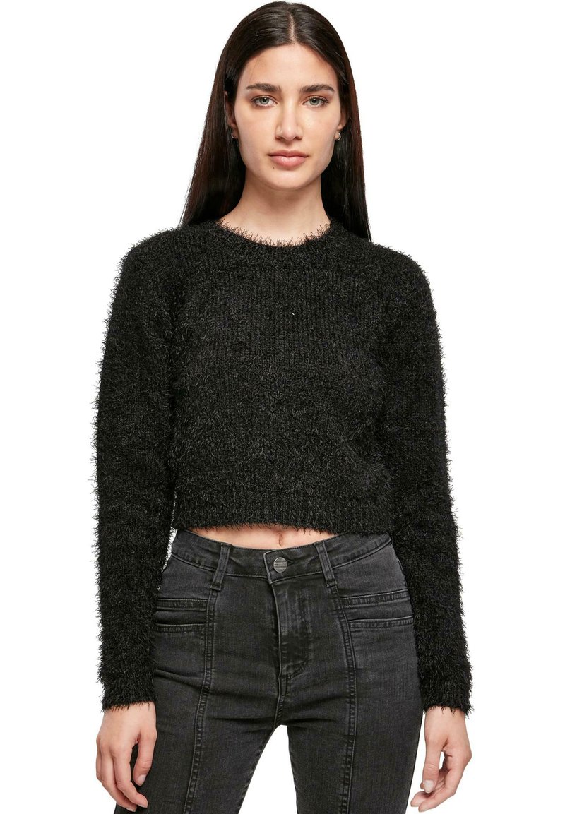 Donna che indossa un maglione corto nera e pelosa, con jeans grigi scuri a vita alta, in piedi davanti a uno sfondo bianco.