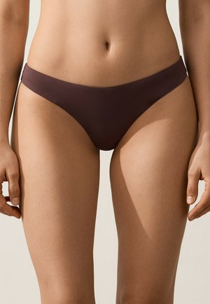 Buste féminin portant un bas de bikini marron foncé, debout avec les bras détendus le long du corps sur un fond uni.