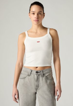Femme portant un débardeur court blanc Levi's et un jean taille haute gris clair, debout devant un fond clair uni.
