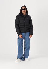 Zwarte pufferjack met een rits aan de voorkant en een staande kraag, gedragen over een grijs shirt. Losse blauwe jeans en witte sneakers. Donkere zonnebril als accessoire.
