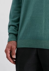 Grüner, leichter Strickpullover mit langen Ärmeln, gerippten Bündchen und Saum. Über schwarzen strukturierten Hosen getragen. Hand ist seitlich sichtbar.