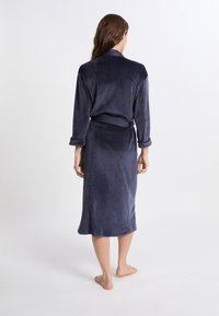 Femme debout, pieds nus, tournant le dos, vêtue d'une longue robe en velours bleu foncé avec des manches retroussées et une ceinture nouée à la taille.