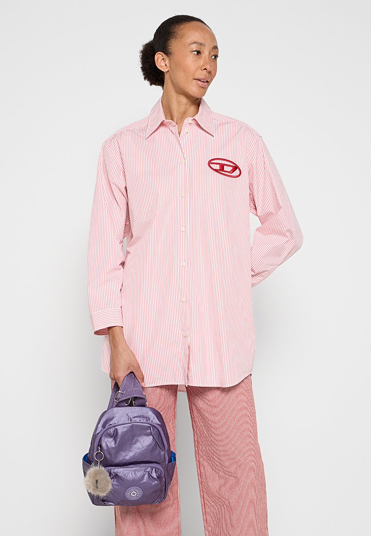 Chemise rayée rouge et blanche avec fermeture à boutons et logo en patch, accompagnée d'un petit sac à dos violet avec un détail en pom-pom en fourrure.