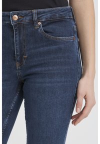 Tuvplāns ar tumši ziliem džinsu biksēm, kas valkātas ar melnu topu, redzama priekšējā kabata, pogas un atslābināta roka, kas atrodas blakus.