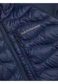 Navy blue insulated jacket with quilted texture, featuring a front zip and “PeakPerformance” logo in silver stitching.