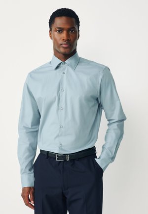 Hombre con una camisa azul claro de manga larga abotonada metida dentro de pantalones oscuros con un cinturón negro, de pie contra un fondo liso.