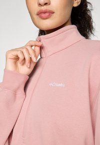 Felpa fleece rosa chiaro con zip, colletto alto, polsini a coste e logo Columbia ricamato in bianco sul petto.