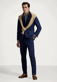 Marineblå dress laget av stoff med glatt tekstur, kombinert med en beige strikkegenser knyttet rundt skuldrene og brune lærsko.