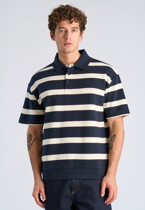 Junger Mann mit lockigem Haar, der ein kurzärmeliges Polohemd mit horizontalen Streifen in Marineblau und Weiß sowie eine dunkle Hose trägt und vor einem einfarbigen Hintergrund steht.