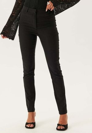 Kobieta w obcisłych czarnych spodniach, przezroczystej czarnej koronkowej bluzce z długim rękawem oraz czarnych sandałkach na obcasie z kokardkami, stojąca na jednolitym tle.