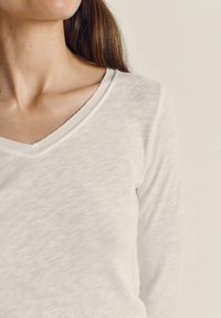 Pruunide pikkade juustega naine, kes kannab valget V-kaelusega pika varrukaga pluusi, kujutatud kaelast kuni keskmise ülakehani heleda tausta vastu.