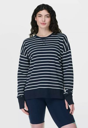 Vrouw die een marineblauw-wit gestreept shirt met lange mouwen en marineblauwe knielange shorts draagt, staand tegen een effen achtergrond.