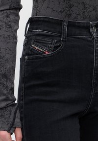 Personne portant un jean en denim noir et une chemise grise à manches longues avec un motif marbré, montrant la zone de la poche avant.