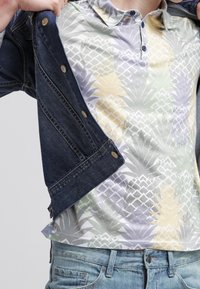 Veste en denim portée sur un polo de couleur claire avec un motif géométrique d'ananas en vert pâle, jaune et lavande. Coupe décontractée et tissu texturé.