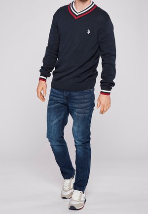 Modèle masculin portant un pull en V bleu marine avec des rayures rouges et blanches, un jean bleu et des baskets blanches, debout devant un fond uni.