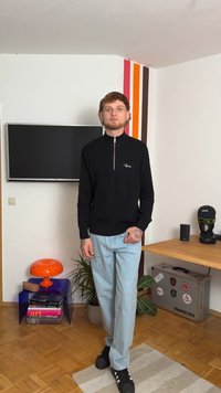 Sweater preto com textura em riscas e fecho éclair, com um pequeno logotipo, calças relaxadas azul claro e sapatos pretos. Lâmpada decorativa em forma de cogumelo laranja ao fundo.