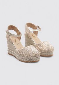 Sandalias de cuña plataforma trenzadas en color beige con una correa, parte superior texturizada y detalle metálico. La suela presenta un diseño de fibra trenzada.