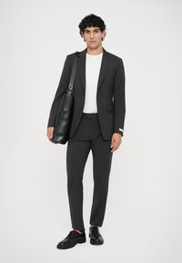 Tmavo šedý oblek so slim-fit nohavicami a jednoradovým sakom, skombinovaný s bielym tričkom a čiernymi koženými topánkami. Čierna taška s ramenným popruhom.
