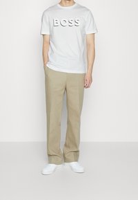 Biały bawełniany t-shirt z napisem „BOSS” w wypukłych czarnych literach, zestawiony z jasnobeżowymi spodniami i białymi trampkami.