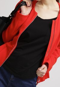 Personne portant une veste rouge ouverte sur un t-shirt noir et tenant une bandoulière de sac à main noir sur son épaule.