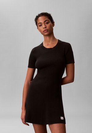 Kvinde med krøllet hår iført en kortærmet, sort ribbestrikket kjole, der står mod en ensfarvet grå baggrund.