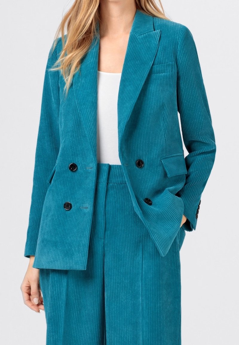 Femme portant un blazer croisé en velours côtelé bleu sarcelle et un pantalon taille haute assorti avec un top blanc en dessous, main dans la poche.